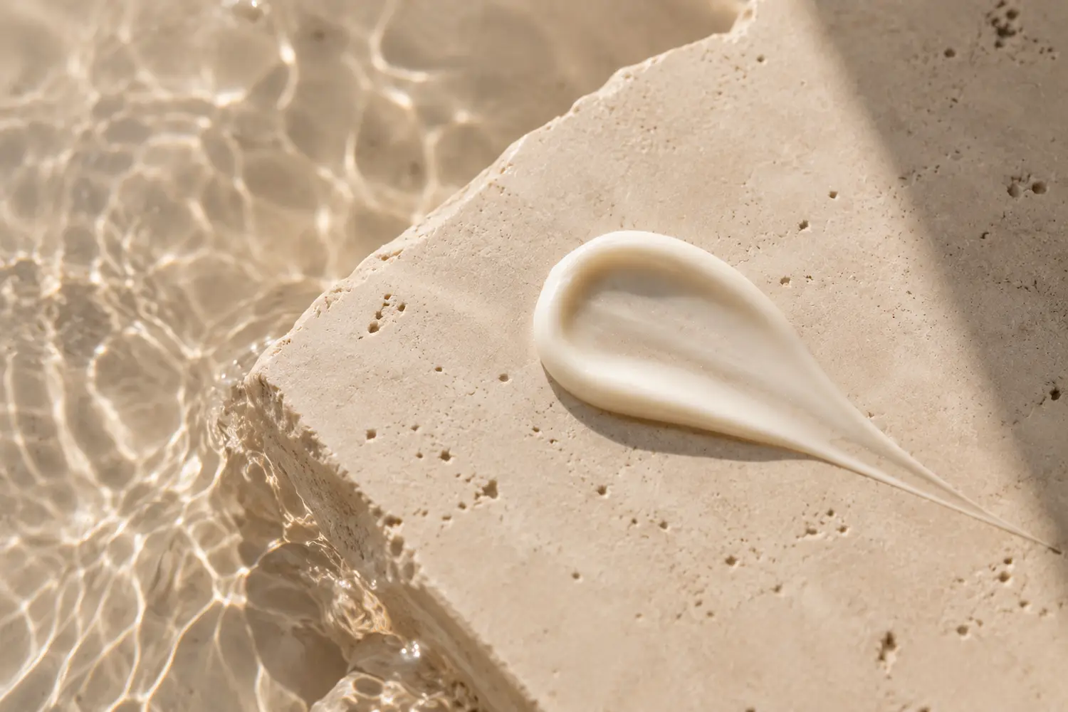 Texture di crema SPF su pietra chiara con riflessi d’acqua e luce naturale in stile beauty editoriale.