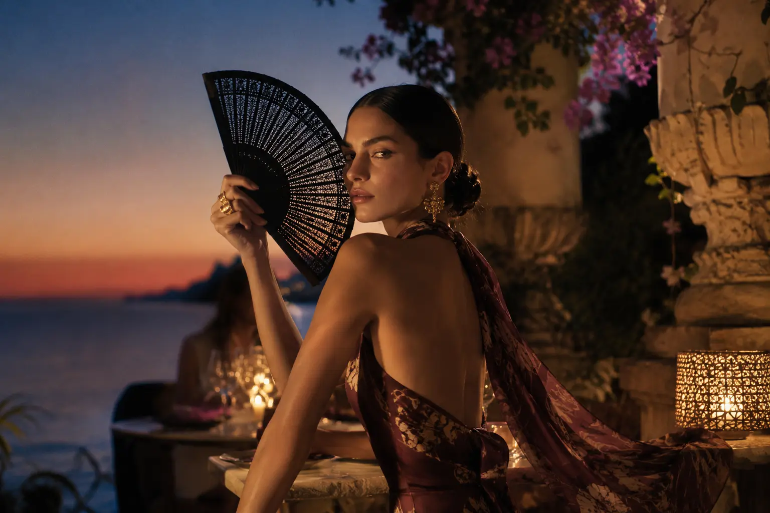 Donna elegante con ventaglio nero durante una cena estiva al tramonto sul mare.