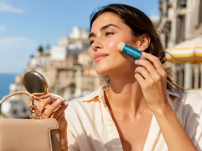 Donna applica Colorescience Brush-On Shield SPF 50 sul viso sopra il make-up durante un ritocco solare all’aperto