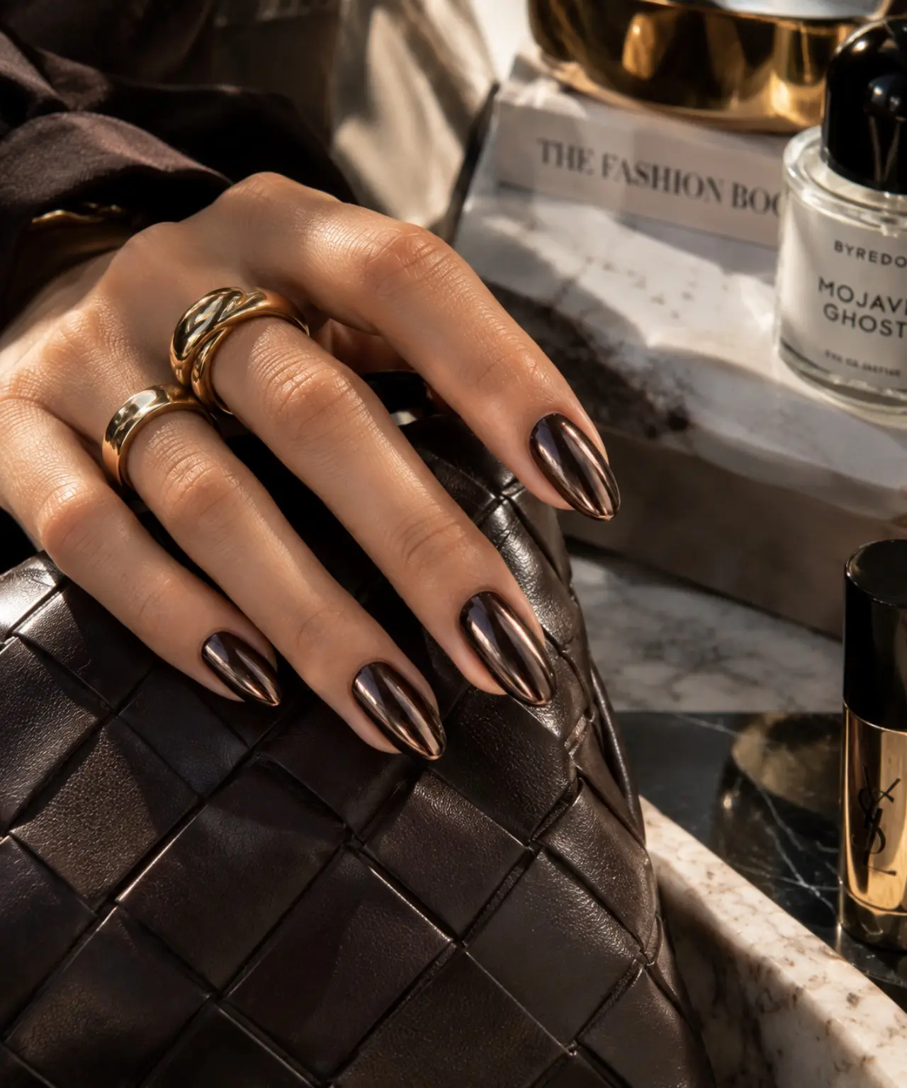 Mano con Dark Chrome Nails bronzo scuro specchiato, riflessi metallici e gioielli oro