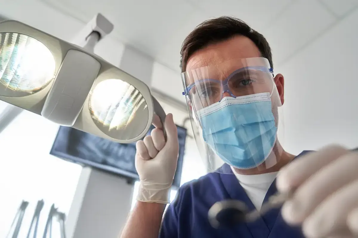 Homem de mascará em um consultório odontológico com um refletor entendendo quanto ganha um dentista recém-formado
