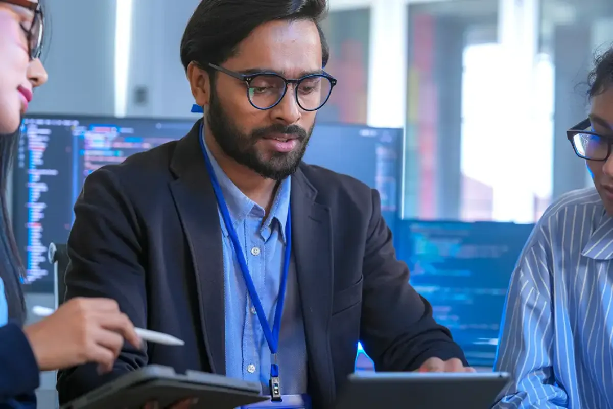 Homem mexendo em tablet com computador ao fundo e 2 pessoas ao lado representando engenharia de software