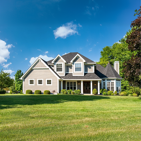 Two-story single-family home with a gray exterior, dark shingle roof, and front porch, set on a wide green lawn with trees under a bright blue sky.
