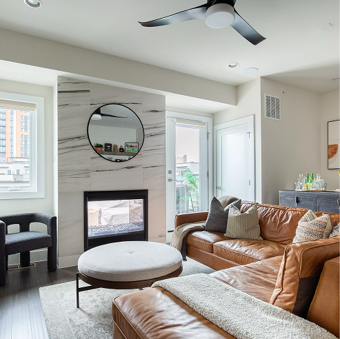 Contemporary living room with a tan leather sectional, round ottoman coffee table, modern ceiling fan, and a marble-accent fireplace with a round mirror above.