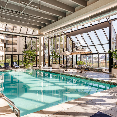 Bright indoor swimming pool in a glass-walled atrium with skylights, modern lounge seating, patio umbrellas, and lush potted plants in a hotel-style amenity space.