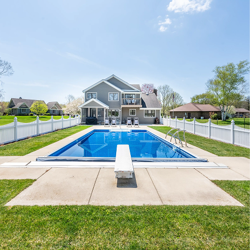 Backyard in-ground swimming pool with a diving board and concrete deck, enclosed by a white fence, with a two-story gray house and patio seating in the background.