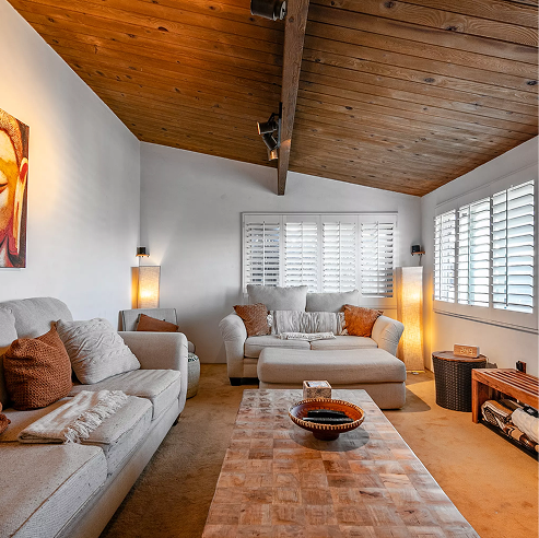 Cozy living room with a vaulted wood ceiling and exposed beam, featuring neutral sofas, a rustic wood coffee table, warm floor lamps, and white plantation shutters.
