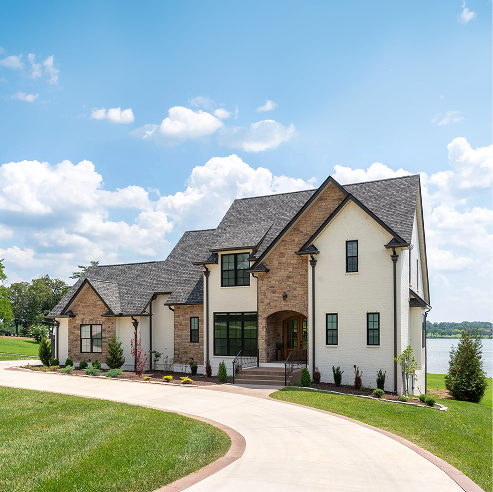 Elegant two-story home with white brick and stone exterior, black-framed windows, and a curved driveway, set on a manicured lawn with a lake view under a blue sky.