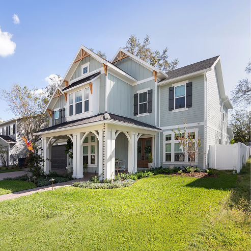 Two-story modern farmhouse-style home with light gray siding, black shutters, and a covered front porch, set on a landscaped lawn in a sunny neighborhood.