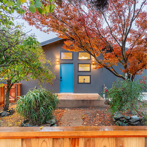 Modern gray home with a bright blue front door and rectangular windows, framed by an autumn tree with orange leaves and a small landscaped front yard.