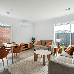 Bright open-plan living and dining room with neutral sofas, wooden coffee table, round dining table with chairs, and large sliding glass doors overlooking a fenced backyard.