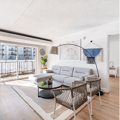 Sunlit modern living room with a gray sectional sofa, black round coffee table, woven accent chairs, and sliding glass doors opening to a balcony with city views.