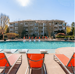 Resort-style outdoor pool with in-water lounge chairs and sun loungers on the deck, set in front of a multi-story apartment community building with landscaped grounds.