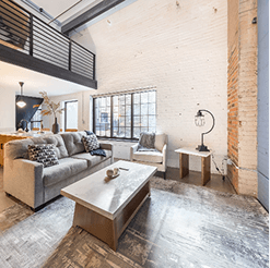 Modern industrial loft living room with exposed white brick walls, mezzanine railing, large factory-style window, gray sofa, armchair, and a minimalist coffee table.