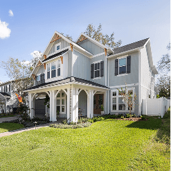 Two-story modern farmhouse-style home with light gray siding, black shutters, and a covered front porch, set on a landscaped lawn in a sunny neighborhood.