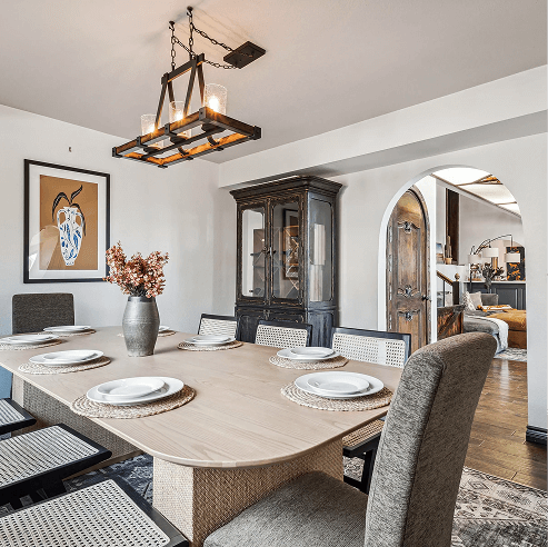 Modern dining room with a light wood table set for six, upholstered chairs, a rectangular lantern-style chandelier, and an arched doorway leading to a cozy living area.