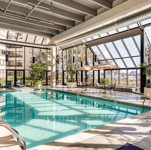 Bright indoor swimming pool in a glass-walled atrium with skylights, modern lounge seating, patio umbrellas, and lush potted plants in a hotel-style amenity space.