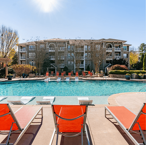 Resort-style outdoor pool with in-water lounge chairs and sun loungers on the deck, set in front of a multi-story apartment community building with landscaped grounds.