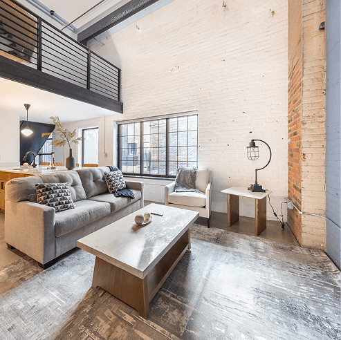 Modern industrial loft living room with exposed white brick walls, mezzanine railing, large factory-style window, gray sofa, armchair, and a minimalist coffee table.