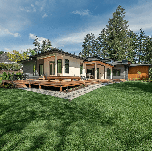 Contemporary single-story home with expansive windows and a wraparound wooden deck, set on a lush green lawn with tall evergreen trees under a clear blue sky.
