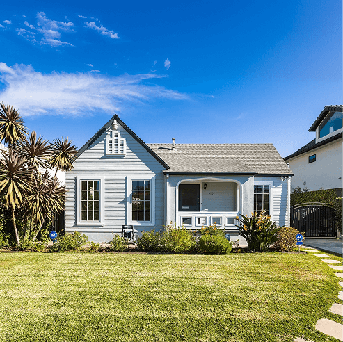 Light-blue cottage with a gabled roof and covered front porch, featuring large windows and a tidy front lawn with drought-tolerant landscaping under a clear blue sky.