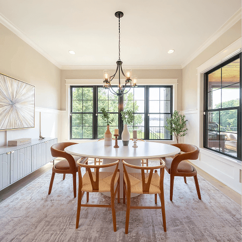 Bright modern dining room with a round white table, four mid-century style chairs, a black chandelier, and large black-framed windows overlooking greenery.