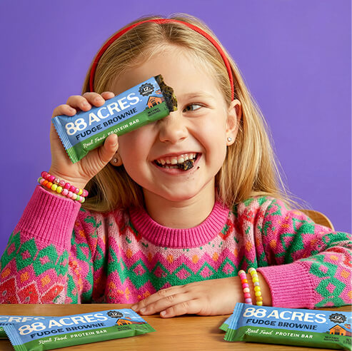 Smiling child holding an 88 Acres fudge brownie protein bar in a colorful lifestyle portrait with product packages on the table.
