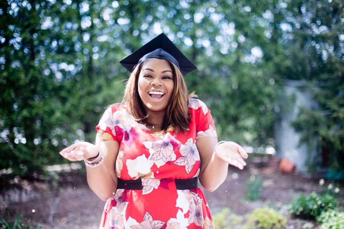 Joyful young woman in a graduation cap and red floral dress shrugging with a big smile in a garden