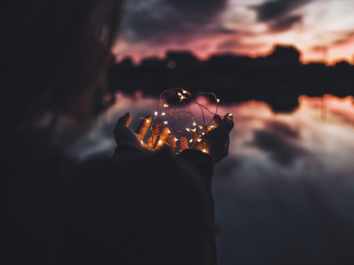 Person silhouetted holding a bundle of glowing fairy lights in cupped hands at sunset by a calm reflective lake
