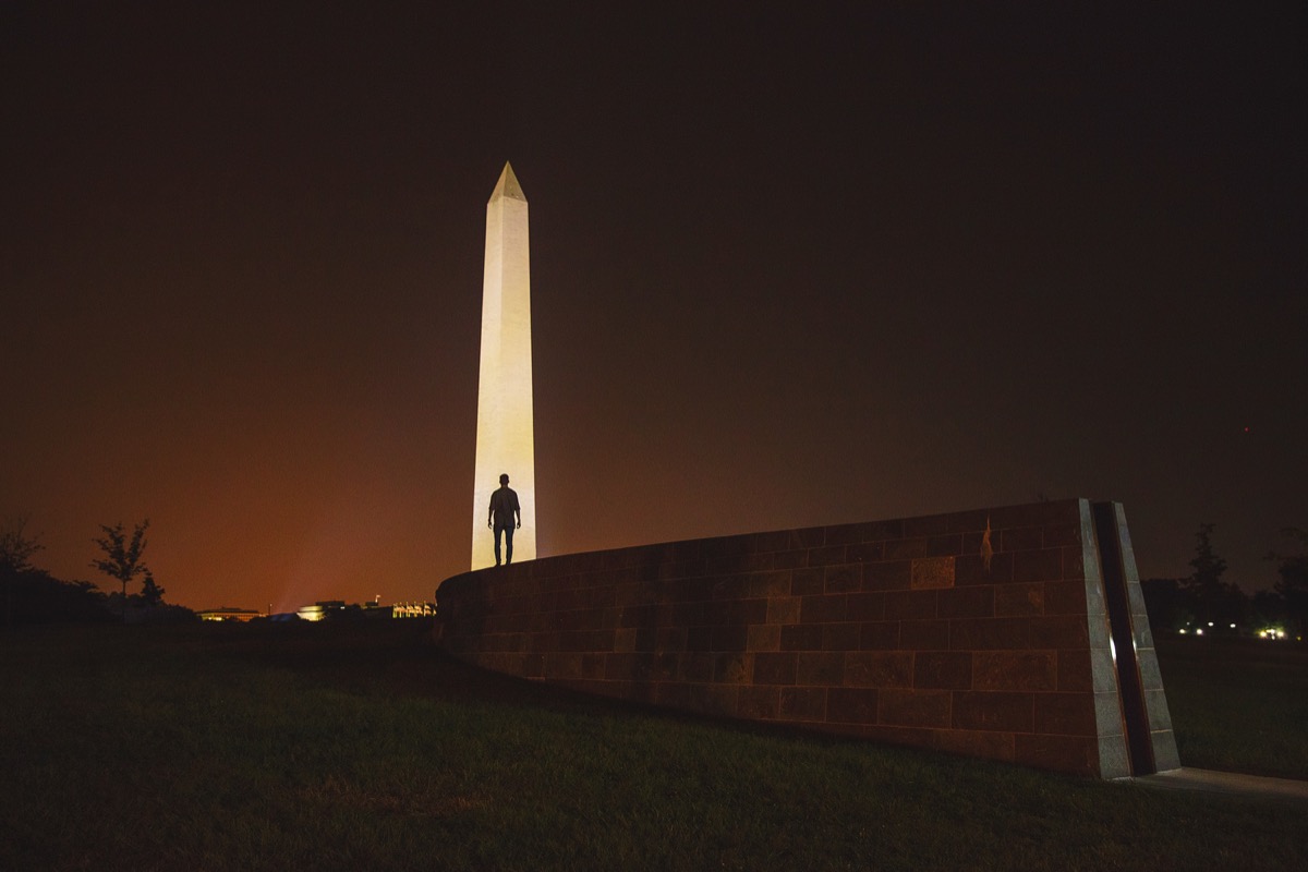 Washington Monument illuminated at night with a small silhouette of a person standing on a stone wall in the foreground