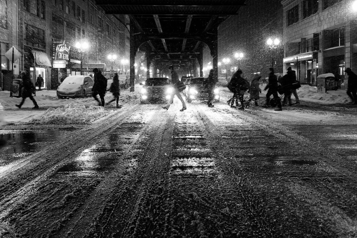 Black-and-white photograph of pedestrians and cars crossing a snowy Chicago street at night beneath the elevated train tracks