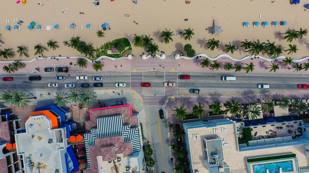 Aerial drone view of a Florida beach showing wavy boardwalk, palm trees, beach umbrellas, road traffic, and rooftops