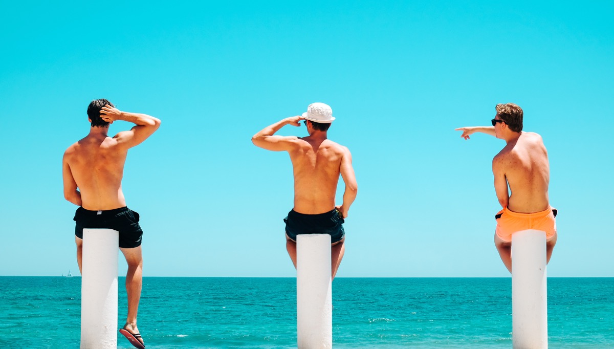 Three shirtless men in swim trunks standing on tall white pillars looking out at the turquoise ocean