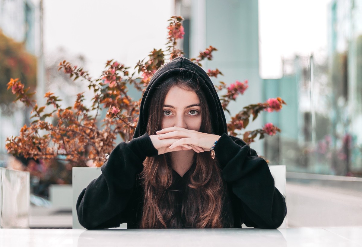 Young woman wearing a black hoodie covering her mouth with her hands, sitting at a café table with bougainvillea flowers behind her