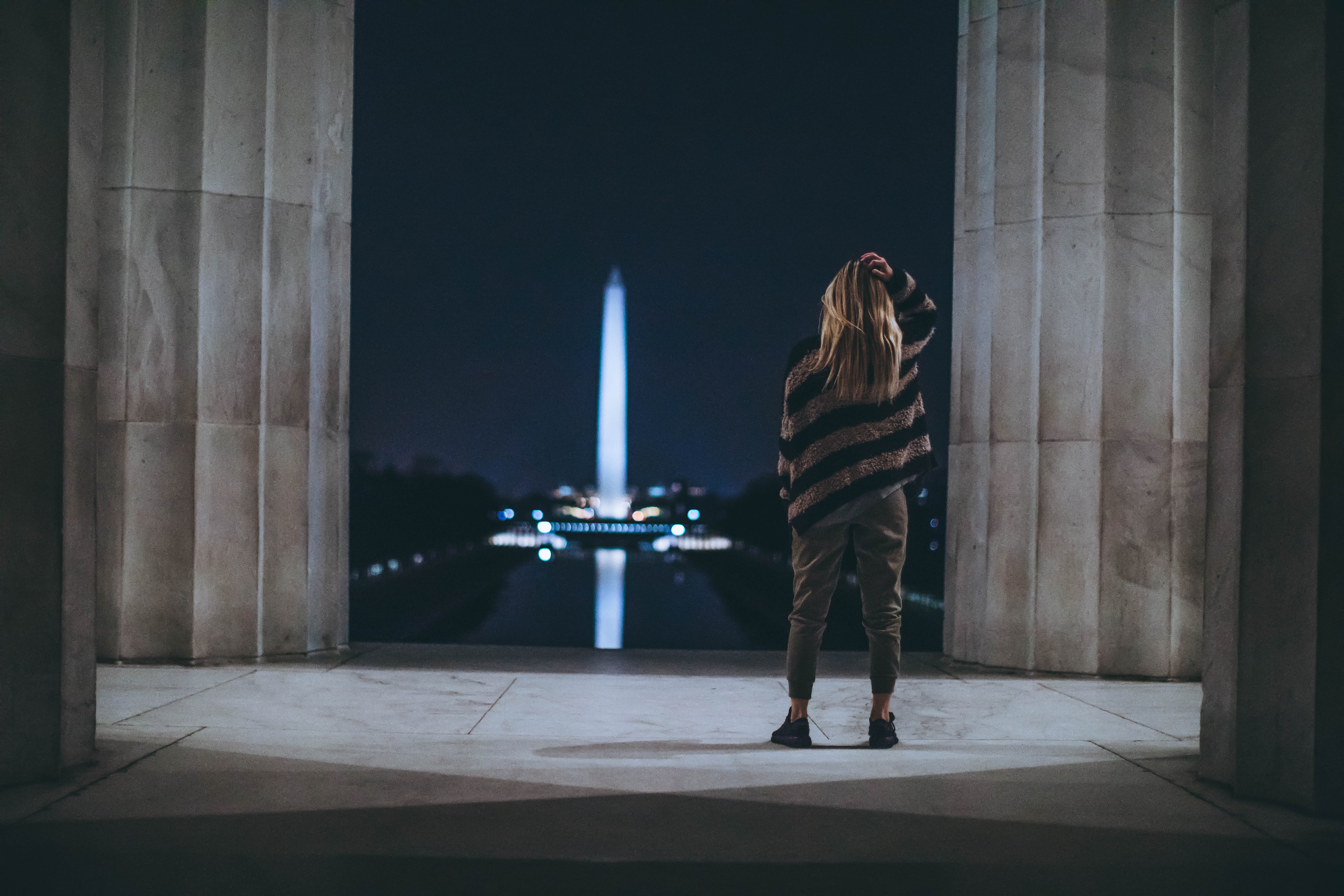 Woman with long blonde hair standing between Lincoln Memorial columns at night looking out at the illuminated Washington Monument and reflecting pool