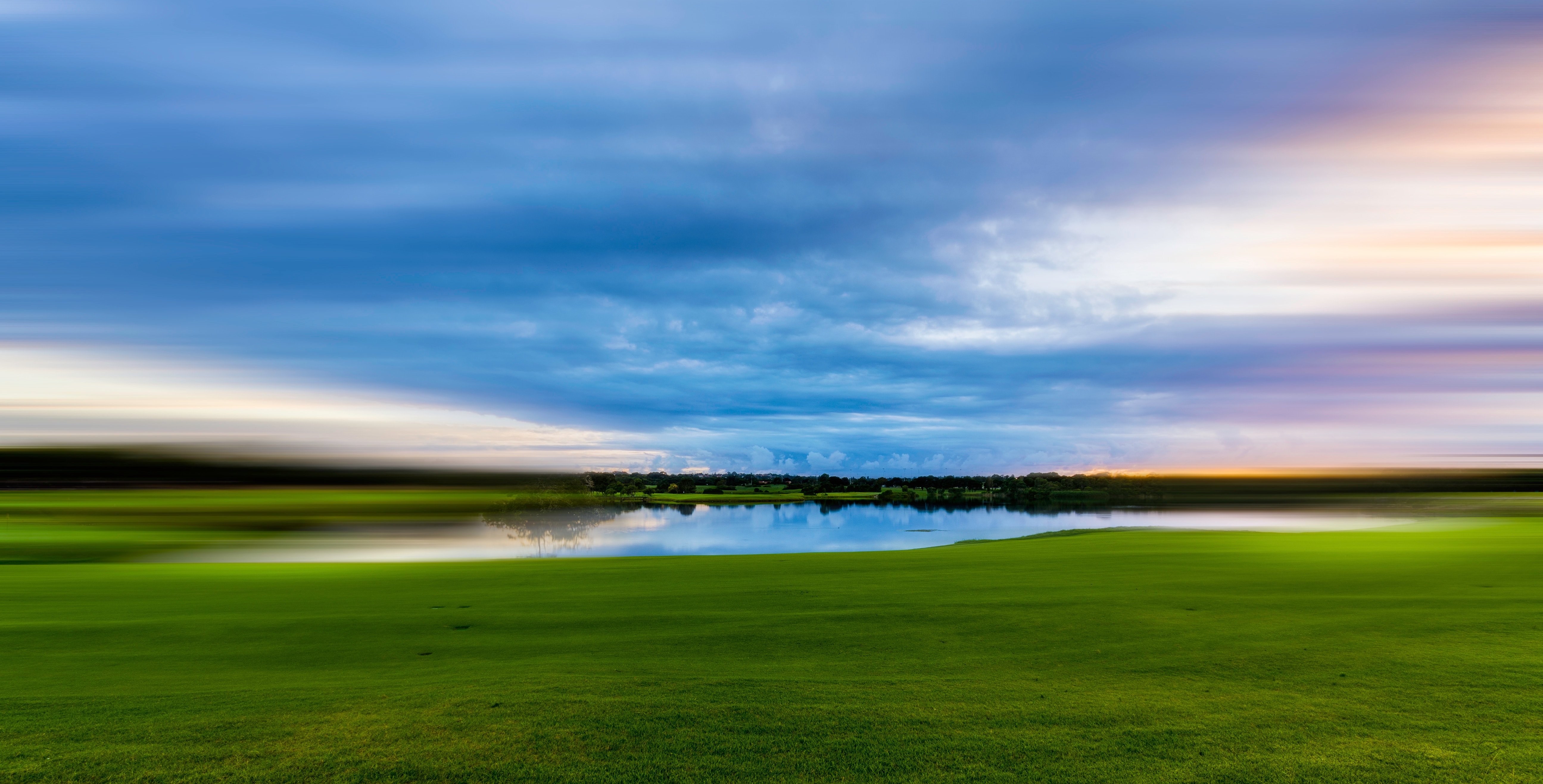 Long-exposure landscape photo of a calm lake at twilight with green grass in the foreground and streaked blue clouds above
