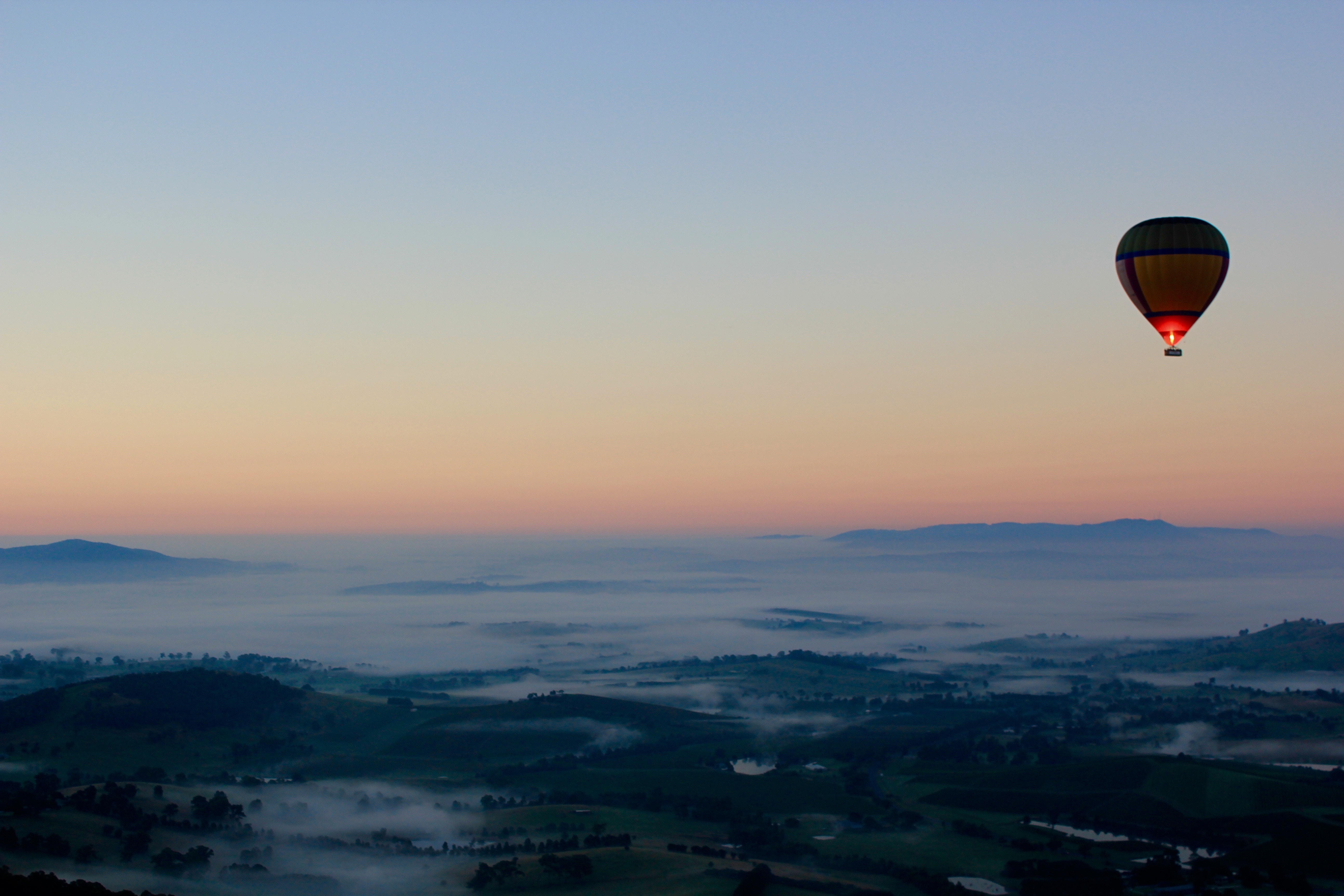Hot air balloon flying over a misty valley at dawn, with rolling hills and a soft pink-orange sky