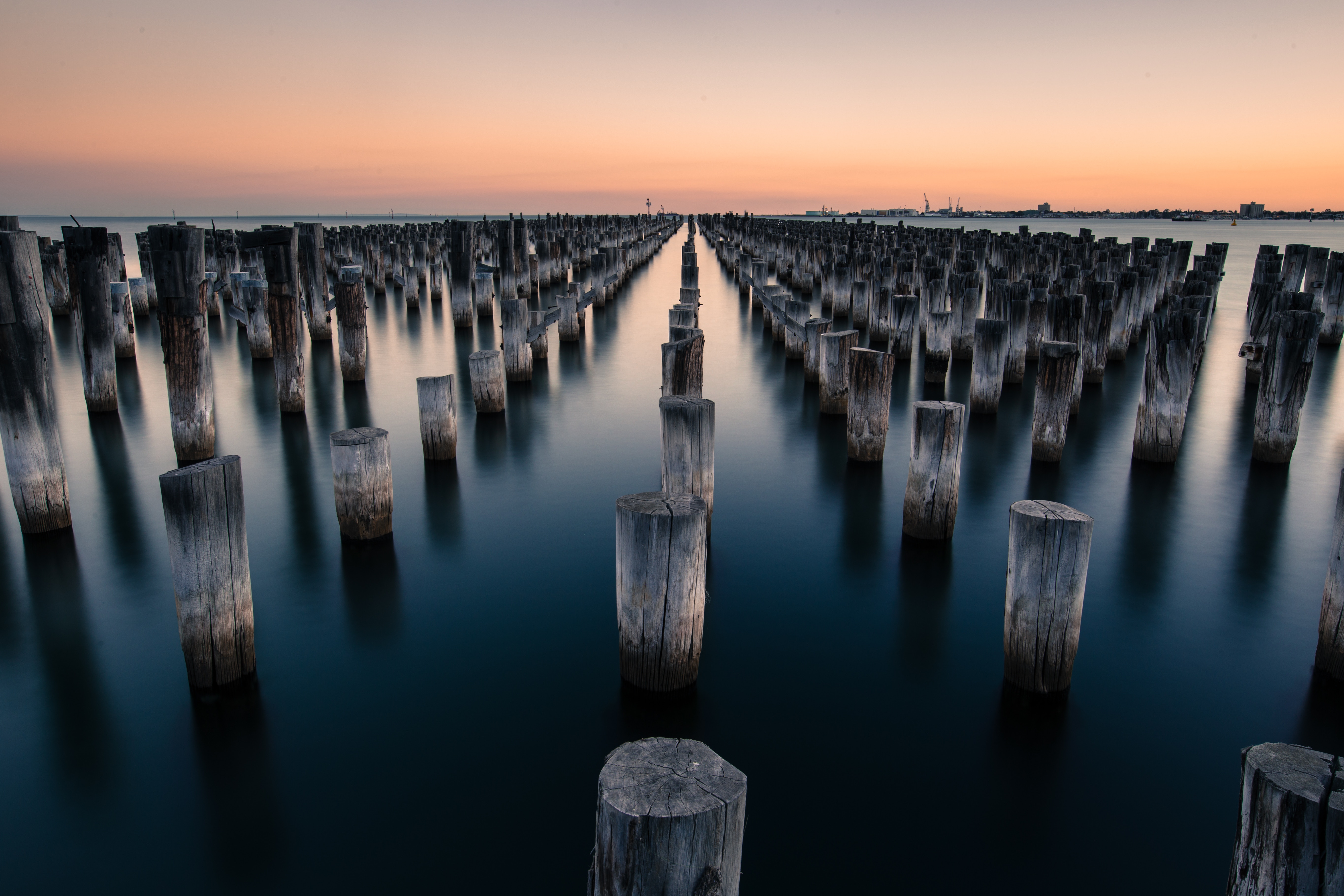 Sunset long-exposure of weathered wooden pilings at Princes Pier in Port Melbourne with calm water reflecting the sky