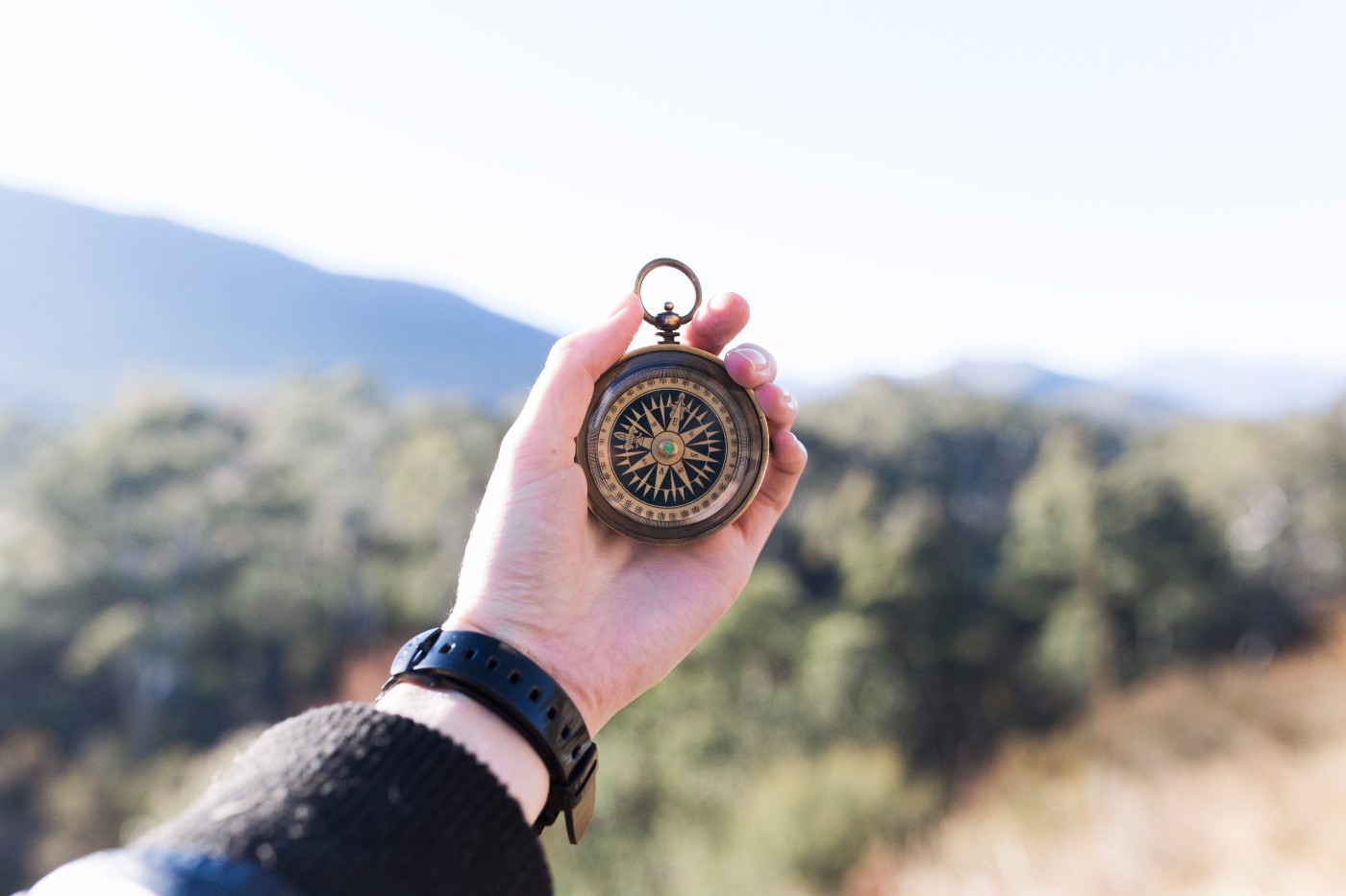Hand wearing a watch holding an antique brass compass against a blurred mountain and forest background