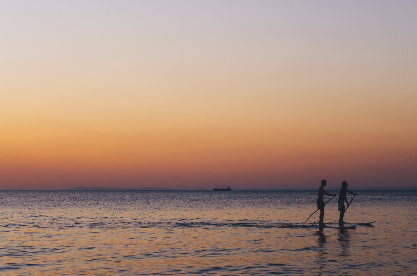 Two silhouetted paddleboarders on calm ocean water at sunset under a peach-colored sky