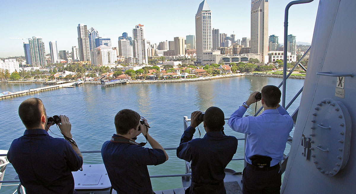 Four US Navy sailors looking through binoculars from a ship's deck toward the San Diego skyline at Seaport Village