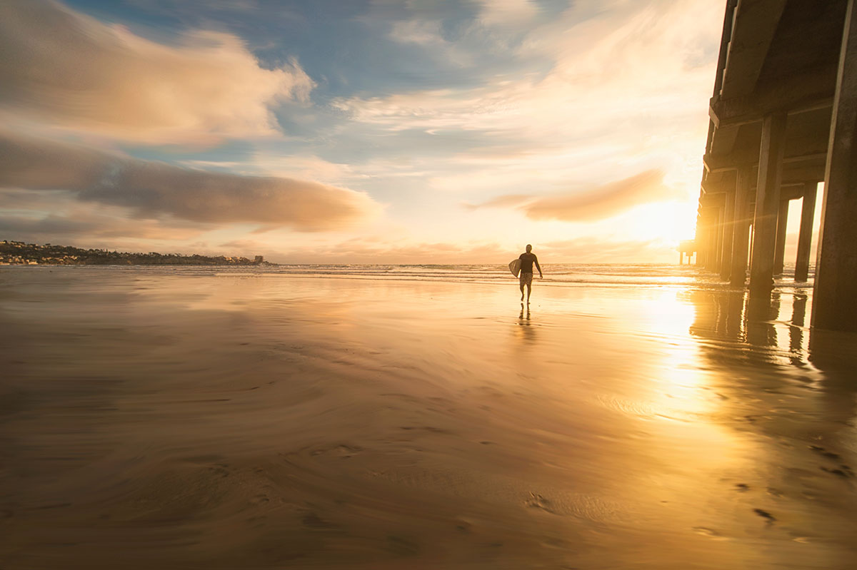 Silhouette of a surfer carrying a board walking on wet sand near Scripps Pier in San Diego at golden sunset