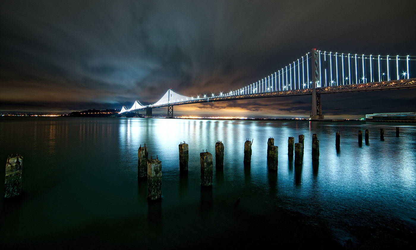 Long-exposure night photo of the San Francisco Bay Bridge illuminated with lights, with old wooden pier pilings in the foreground