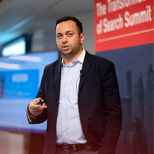 Man in a suit presenting at the Search Summit conference on a bright stage.