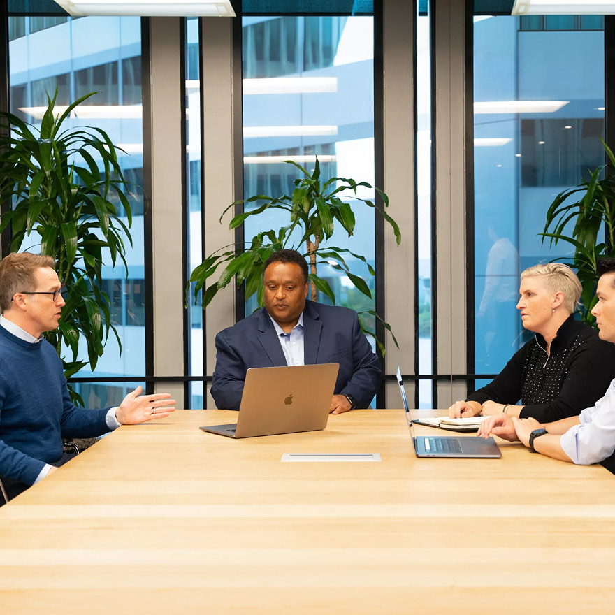 Corporate team collaborating in a meeting inside a modern glass conference room with laptops.