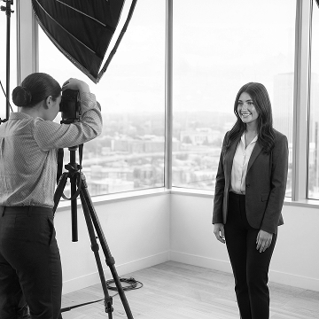 Photographer on a tripod capturing a woman in an office with a city view behind her.
