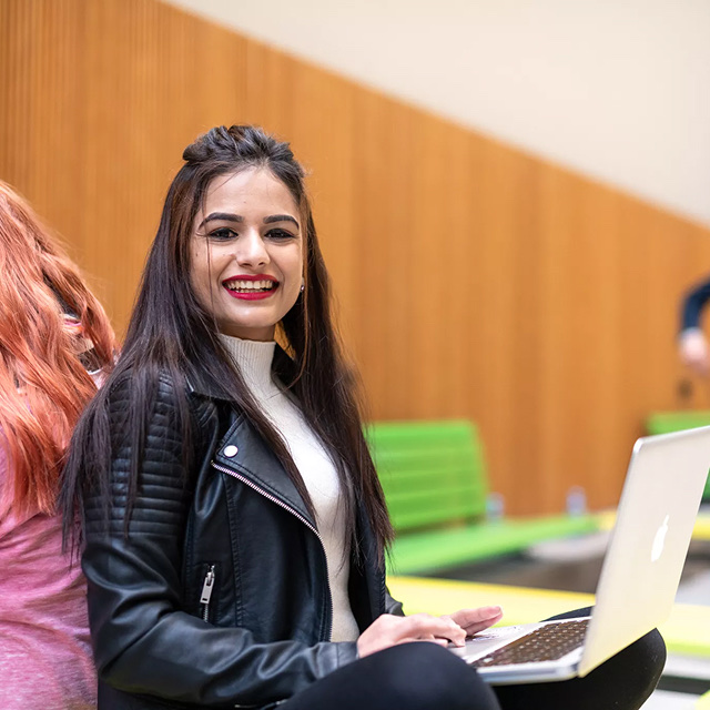 Young woman smiling at her laptop while seated in an open university event venue space.