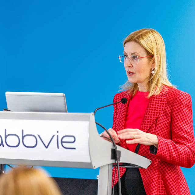 Woman in a red jacket speaking at a podium during an AbbVie company conference event.