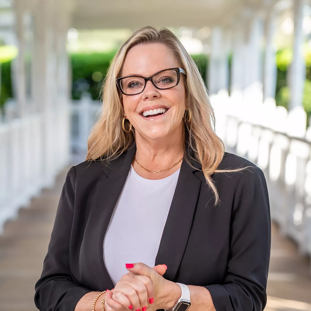 Blonde woman wearing glasses and a blazer laughing naturally in an outdoor professional portrait.