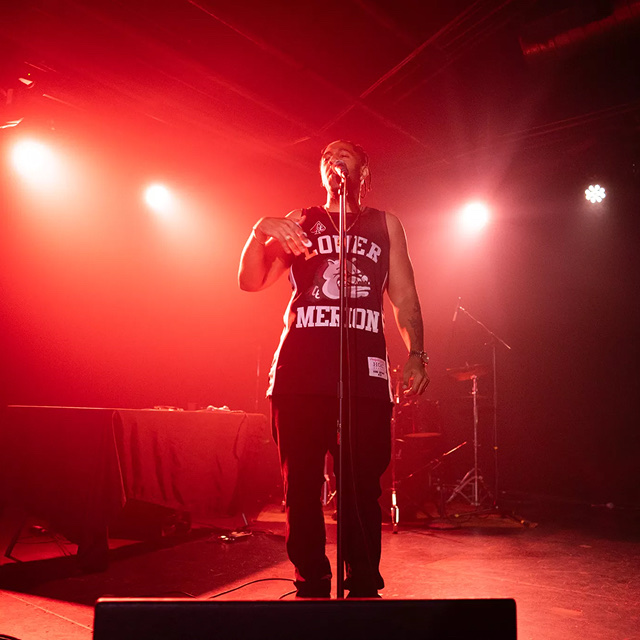 Hip-hop artist performing with energy on stage bathed in red concert lighting.